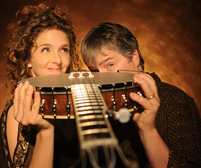 Bela Fleck and Abigail Washburn pose, peering over a banjo in their hands on a brown background